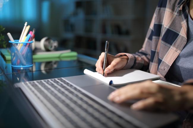 Bilde av hender som skriver på en blokk med en PC ved siden av