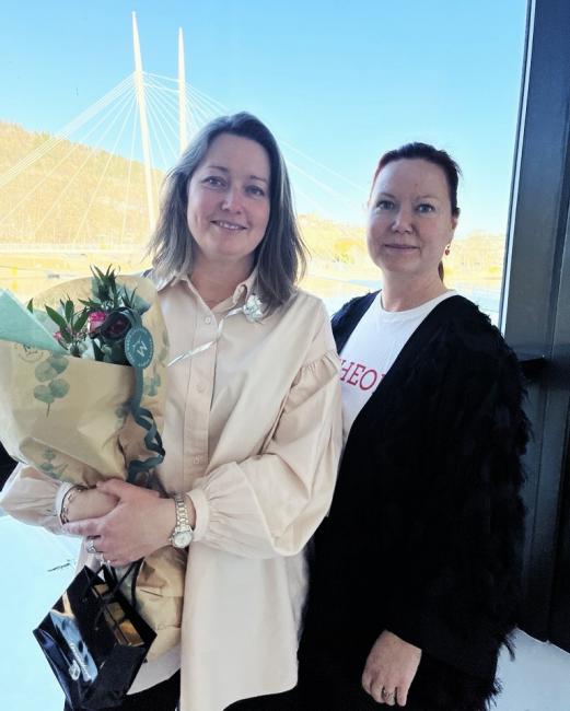 Bilde av mørkeblond smilende dame med blomsterbukett og en annen, mer alvorlig dame med Drammenselva i bakgrunnen.
